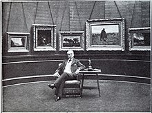 Henry Vasnier est assis sur une chaise, accoudé à une table, devant un mur de sa galerie où sont accrochés plusieurs de ses tableaux.