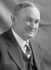 Een man met dunner wordend peper-en-zoutkleurig haar, gekleed in een zwarte jas en vest, een wit overhemd en een stropdas met patroon