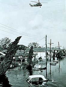 Monochrome image of a flooded street. Cars are submerged, and a helicopter is visible hovering overhead.