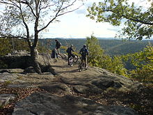 Fotografía de excursionistas y ciclistas de montaña en la cima de una colina de roca plana con vistas a un bosque