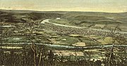 A tinted postcard depicts a town as seen from a hillside. It is laid out on a triangle of flat land between two converging streams. The larger and more distant of the two streams is flowing from mountains, while the smaller stream flows through farmland.