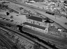 Eine Schwarz-Weiß-Luftaufnahme eines zweistöckigen Bahnhofs.Eine einzelne Spur auf einem Damm gegenüber dem Hauptbahnhof hat einen kleinen Unterstand.