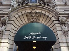 A entrada para o Ansonia.  Um toldo verde com as palavras "The Ansonia, 2109 Broadway" é colocado no topo da porta.  A entrada é cercada por blocos de calcário e há uma face de calcário esculpida acima da entrada.