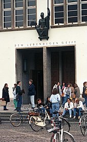 L'entrée d'un bâtiment blanc devant laquelle discutent des étudiants. Au premier plan, des vélos.