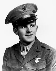 Askeri elbise üniforması giyen genç bir adamın siyah beyaz görüntüsü. Şapka takıyor ve yukarı bakıyor ve gülümsüyor. Tüfek nişancılık rozeti, üniformasının sol göğsünde açıkça görülüyor.