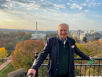 Carleton Varney in a blue suit, green vest, and pink pocket square stands on a balcony which overlooks the White House and Washington Monument surrounded by the changing autumnal trees.