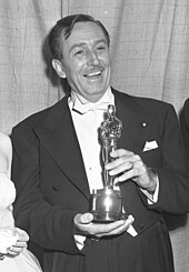 Uma fotografia em preto e branco de Walt Disney em pé, segurando um Oscar.