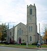 A light gray stone building seen from across a street with a pointed roof in the middle, a tower on the left and two sets of pointed-arch red doors in the center and right. A banner on the left of the center door says "Celebration" in vertical type.