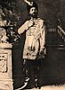 Photograph of a man standing, wearing leather books, knee-length tunic and feathered hat