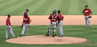 Seven men wearing red baseball jerseys, gray pants, and black caps are standing on a baseball diamond's infield; three are talking together on the mound.