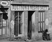 George N. Barnard's photograph of a slave trader's business on Whitehall Street Atlanta, Georgia, 1864. A United States Colored Troop Infantry corporal is sitting by the door. United States Colored Troop enlisted African-American soldier reading at 8 Whitehall Street, Atlanta slave auction house, Fall 1864- 'Auction & Negro Sales,' Whitehall Street LOC cwpb.03351 (cropped).tif