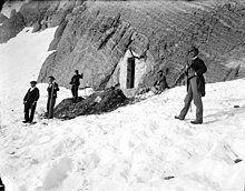 Photographie d'un groupe d'hommes rassemblés sur la neige devant l'entrée d'une grotte.