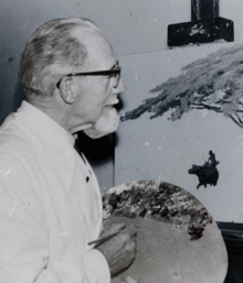 Waist up view of a grey haired man with white mustache and beard and wearing glasses facing right with pallet in his left hand and paint brush in his right hand stands at a paint easel holding an unfinished painting