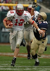 Ein American-Football-Spieler, der den Ball in einer weißen Uniform mit roter Schrift hält, weicht einem Tackle eines anderen Spielers in einer blauen Uniform mit weißer Schrift aus