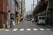 English: A street scene in Akihabara. The image shows several people and cars, buildings to the left and the train lines that leave Akihabara Station to the north