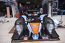 Photographie d'une voiture de sport-prototype bleu, noir et orange, en pièces détachées, vue de face, sur une allée.