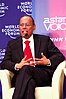 A middle-aged Indian man wearing a black suit, white shirt and red tie speaking into a microphone.