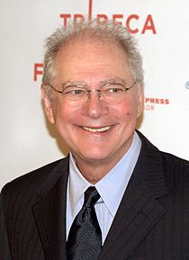 A white-haired man wearing glasses and a suit smiles and looks off-screen.