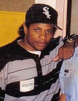 Rapper Eazy-E usando uma camisa polo listrada e um boné preto com o logo na frente do time de baseball Chicago White Sox na cor branca.