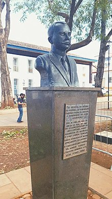 Na imagem há uma estatua, de cor cinza escuro, do busto de um homem. Abaixo do busto há uma placa.