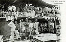 Deux hommes blancs en tenue coloniale et un groupe d'hommes noirs devant un bateau