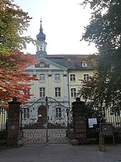 Bâtiment baroque avec un fronton et un clocheton, peint en jaune clair derrière une grille de fer forgé.