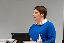 A short-haired brunette woman in a blue sweater stands at a podium and speaks