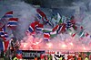 A section of a stadium with supporters waving flags and holding flares which have created a lot of smoke