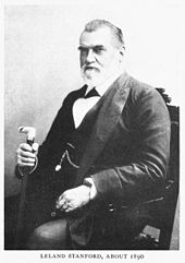 Elderly Caucasian male, wearing a formal suit, with a watch fob, sitting in a chair and holding a cane.