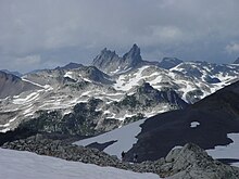 Lightly glaciated mountainous landscape with a steep vertical rocky mountain towering above the surrounding terrain in the background.