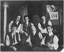 A black and white photograph of six young-to-middle-aged men, all wearing white shirts, dark ties and waistcoats.