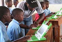 Close-up photograph of schoolchildren in a classroom with green laptops, being assisted by an adult