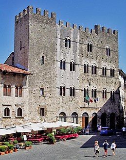 Внушительное четырехэтажное каменное здание с бойницами и рядами спаренных окон, выходящее на городскую площадь.