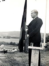 Un homme debout en extérieur derrière un pupitre en bois. Derrière lui, un homme se tient près d'un immense drapeau norvégien.