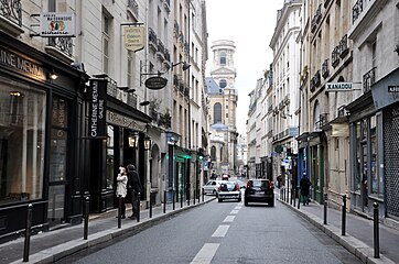 サン＝シュルピス通りをサン＝シュルピス広場方向に見る。背景はサン＝シュルピス教会 (Rue Saint-Sulpice en direction de la place Saint-Sulpice ; en arrière-plan, la tour nord de l'église Saint-Sulpice de Paris.)