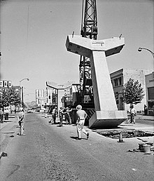 Construction crews look on as a mobile crane hoists a Y-shaped concrete column into the middle of a city street.