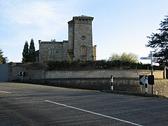 Stone building with tower to right hand side. In front is a wall separating the building from the road.