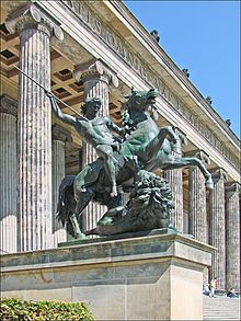 Sculpture of a nude man astride a horse. He aims a spear at an attacking lion.