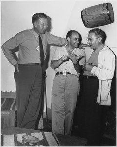 Three men talking. The one on the left is wearing a tie and leans against a wall. He stands with his head and shoulders visibly above the other two's heads. The one in the center is smiling, and wearing an open-necked shirt. The one on the right wears a shirt and lab coat. All three have photo ID passes.