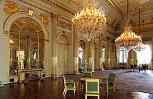 Photographie en couleur d'une vaste salle avec du parquet, des colonnades dorées, un plafond chargé de dorures, des lustres, une petite table et quatre fauteuils verts.