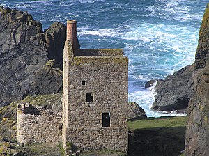 A small stone building obstructs a view of waves crashing into rocks behind it.