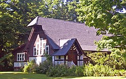 A brown and white building with high, steep roof hanging over at one end in the middle of some trees