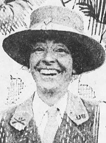 زنی سفید پوست ، که لبخند گشاد می زند ، کلاهی لبه دار ، یقه گردن و ژاکت یکنواخت به سر دارد.