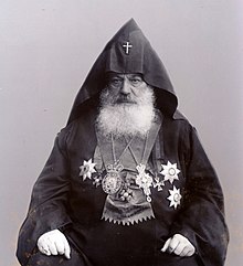 Fénykép egy fehér szakállú, ülő és örmény hivatali öltözetet viselő férfiről.