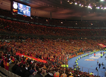 Photo de spectateurs, habillés en dominante rouge et jaune, dans une tribune d'un stade de football. Un écran géant est visible.