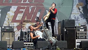 A wide shot with speaker boxes and stage equipment to either side of Owen, Strachan and Cheney. Owen is down on his left knee holding his double bass with his left hand high on the neck and bracing it on his right knee. He has a tattoo visible on his upper, inner left arm. Strachan is partly obscured behind his kit and wears a hat. Cheney is high on the left side of the bass with his left leg at mid-way and right leg at the start of the neck. He plays his electric guitar with his left arm on its neck and right arm with a pick near the strings. The stage backdrop includes the stylised lettering of the band's name with only 'End' within shot.