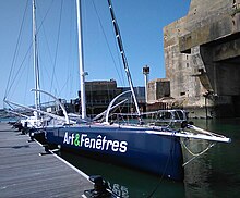 Vu de trois quarts avant, voilier noir avec foils en C, amarré au ponton.