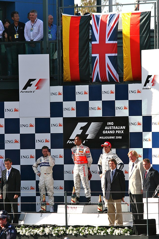 Three men in racing overalls standing on the podium with smartly dressed officials to their right and left