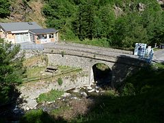 Fotografía en color de un puente de piedra que atraviesa un arroyo de montaña.  Al final del puente, a la izquierda de la foto, edificios recientes de una sola planta.