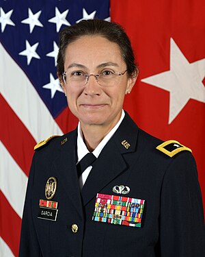 A photograph of a female American military officer in uniform, in front of the flag of the United States.